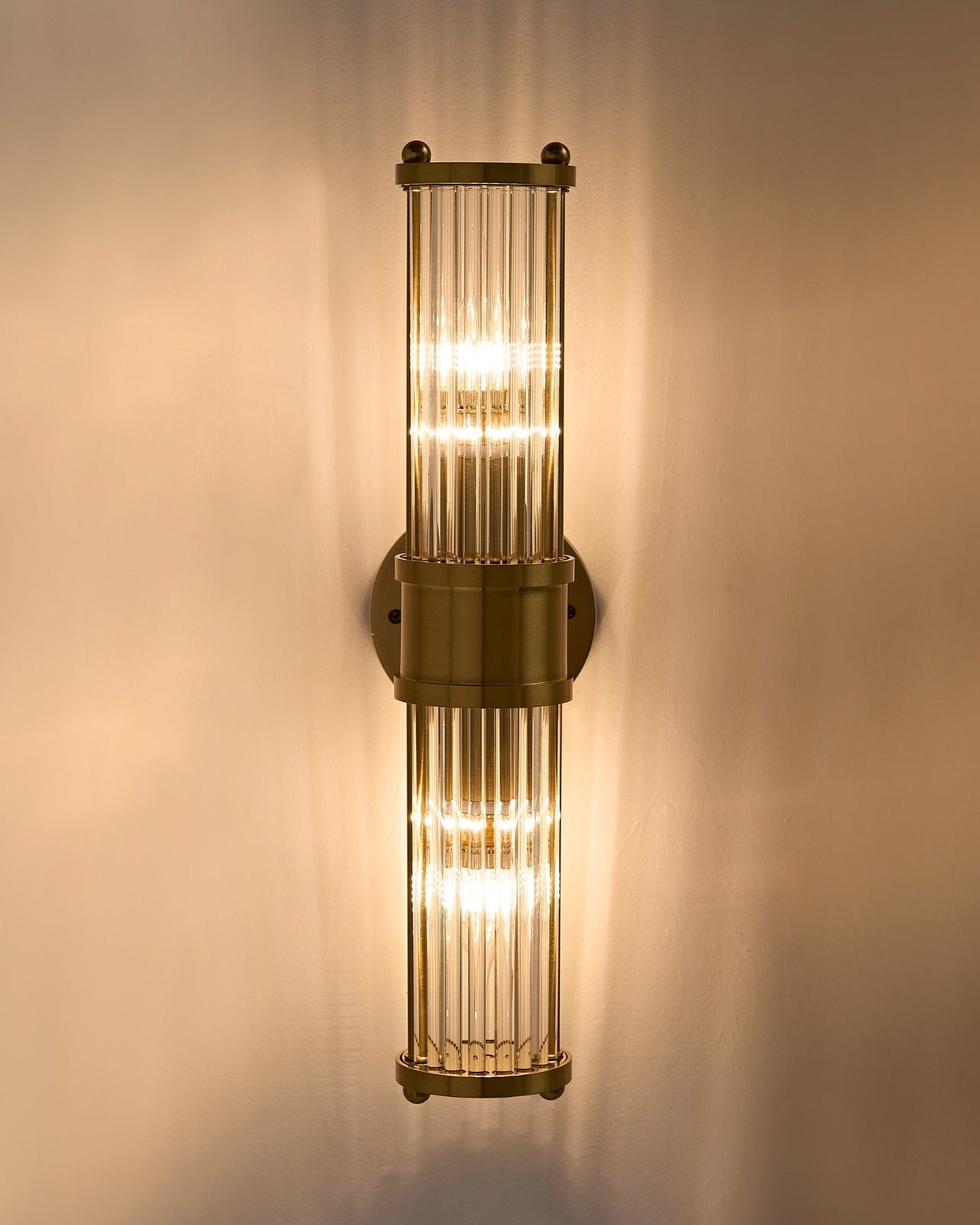 Brimsley II Wall Sconce illuminated on the wall, featuring a vertical brass frame with ripple-textured clear glass panels casting a warm, ambient glow upward and downward.