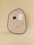 Asymmetrical fabric-covered pinboard with a brown outer frame, featuring pinned Pantone color cards, a small sketch, and gold jewelry.