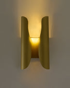 Sebastian wall sconce in brushed brass, illuminated on a wall, featuring a sculptural curved double-panel design that casts a warm, ambient glow upward and downward.