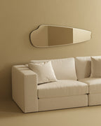 Horizontally mounted frameless wall mirror with an elongated organic shape, displayed above a light-colored sofa in a minimalist interior.
