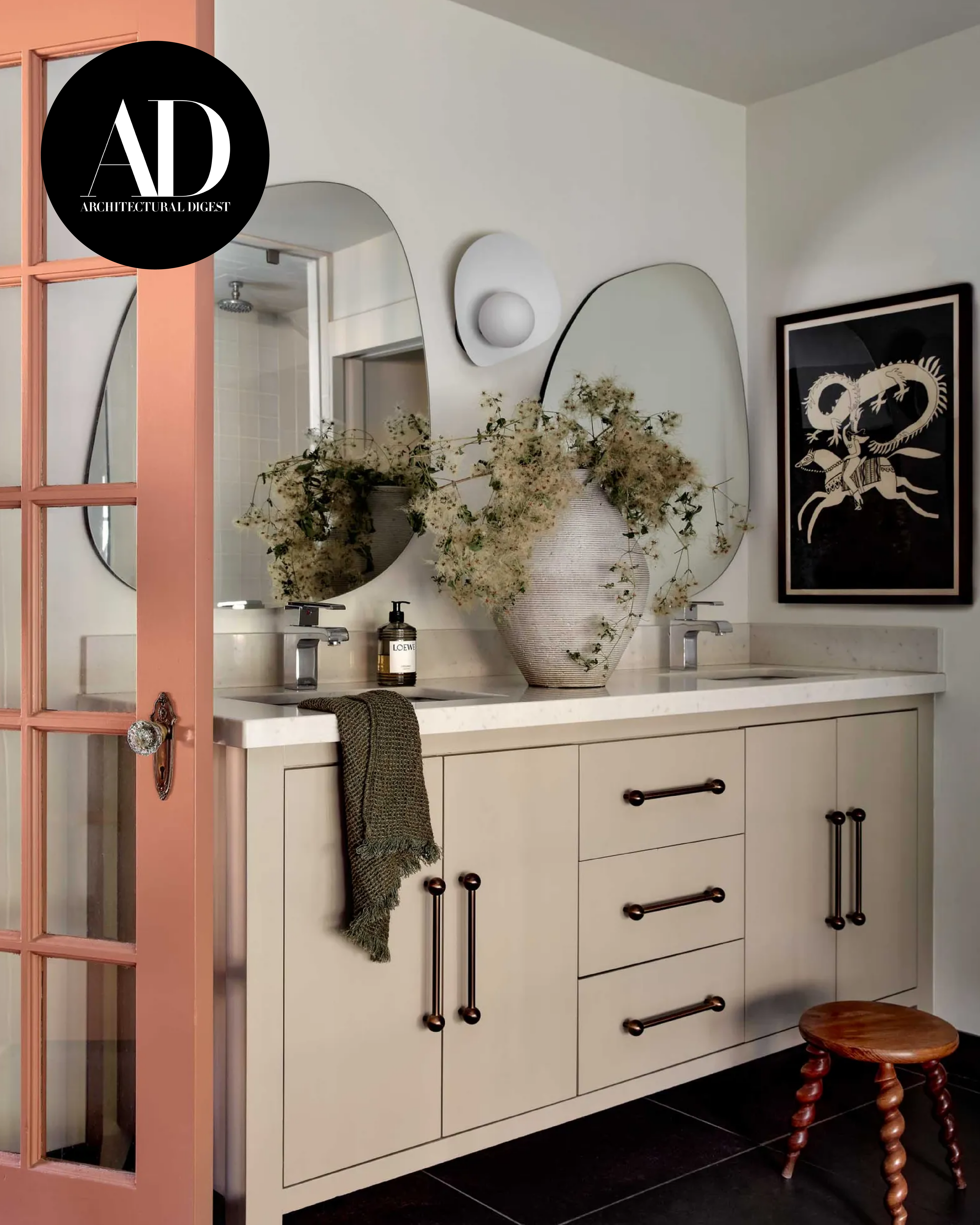 Architectural Digest–featured bathroom vanity with double mirrors, light cabinetry, a ceramic vessel with dried florals, and a soft pink door frame.