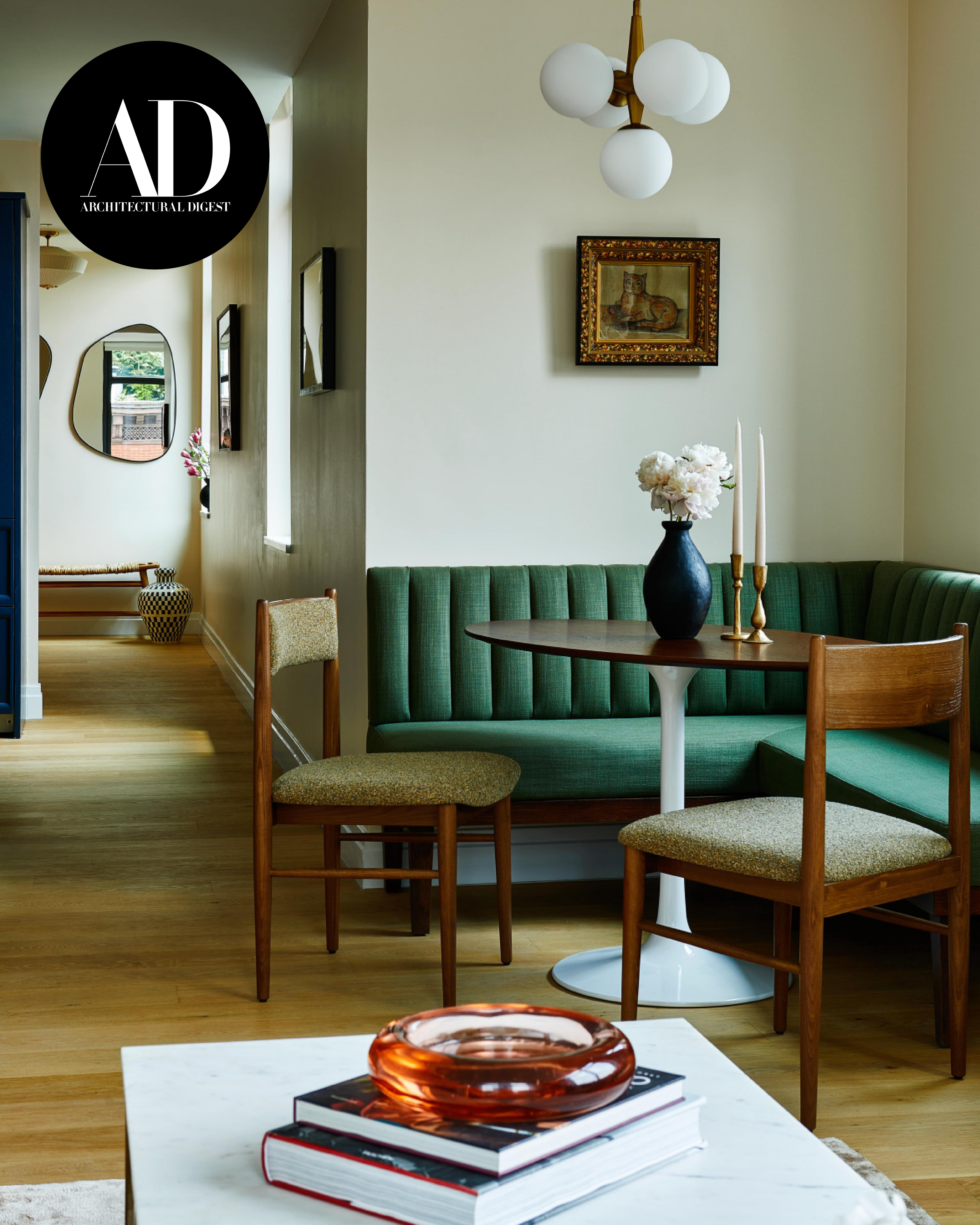 Architectural Digest interior feature showing a green upholstered banquette, round dining table, wooden chairs, and asymmetrical wall mirrors in a softly lit home.