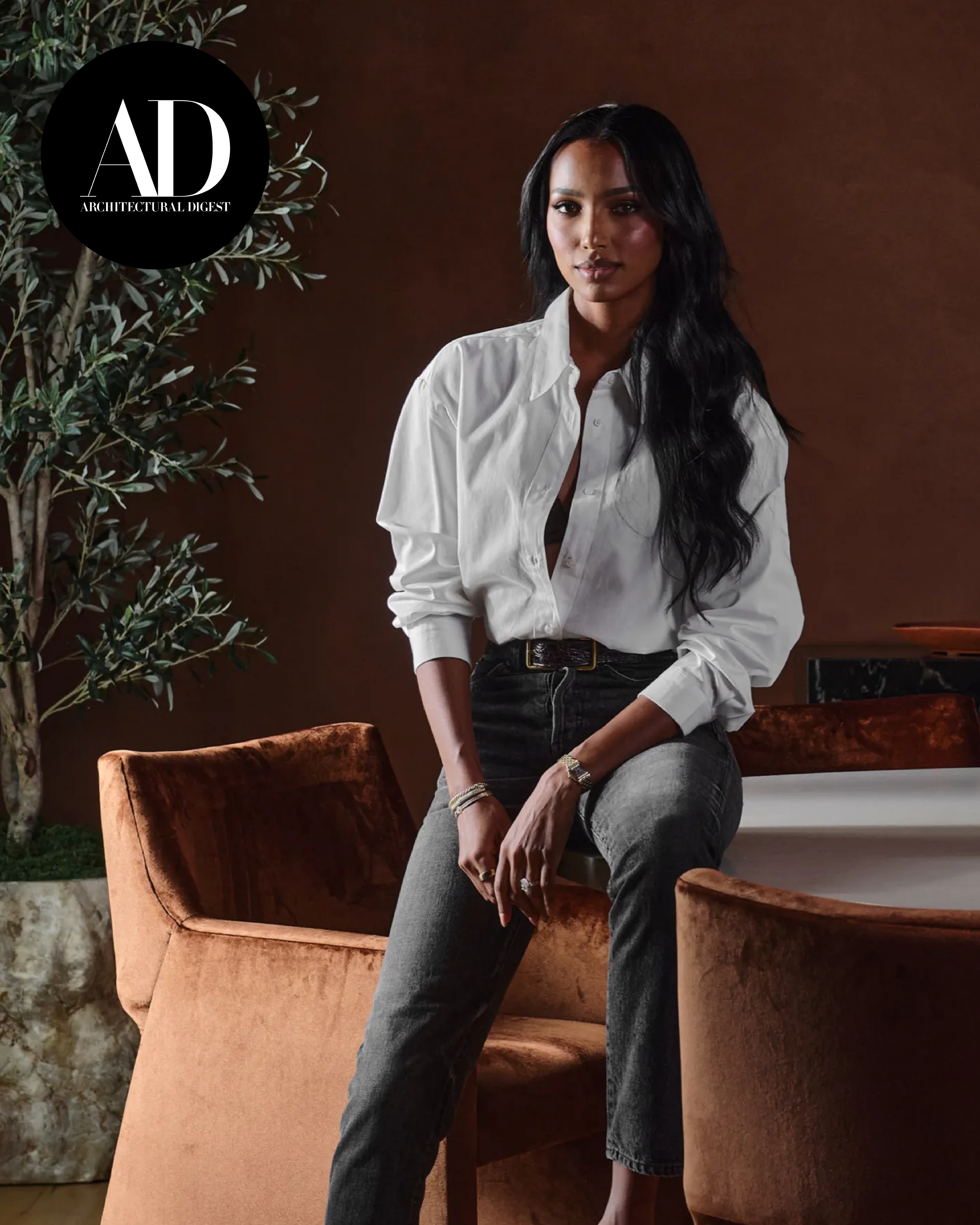 Architectural Digest portrait showing Jasmine Tookes seated on a velvet chair in a softly lit interior with warm brown walls, indoor plants, and contemporary furniture.