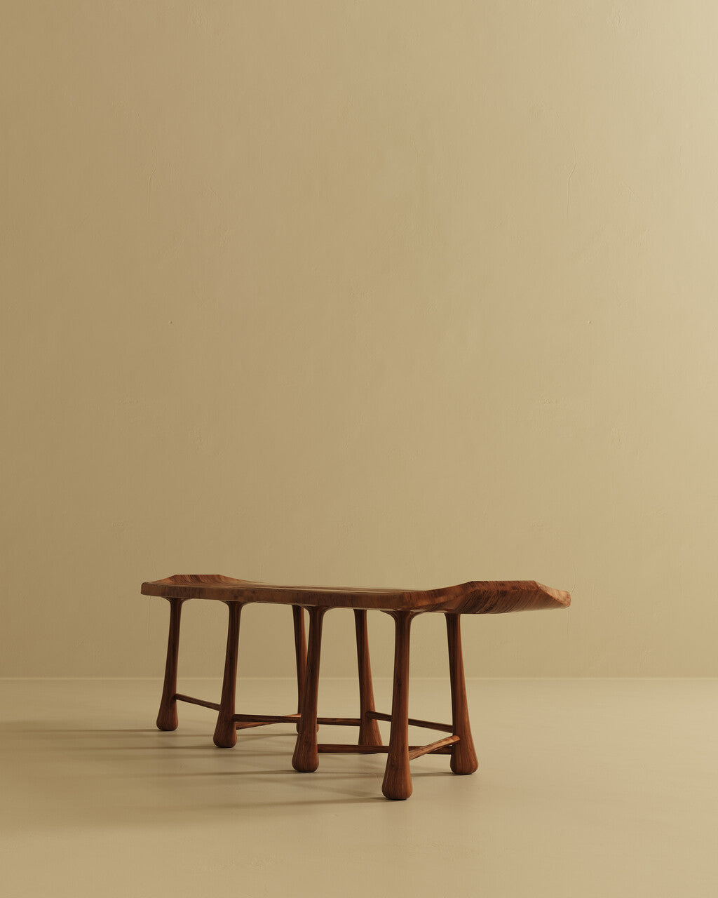 A long wooden bench with a flat seat and multiple rounded legs, shown from a low angled perspective against a plain wall and floor.