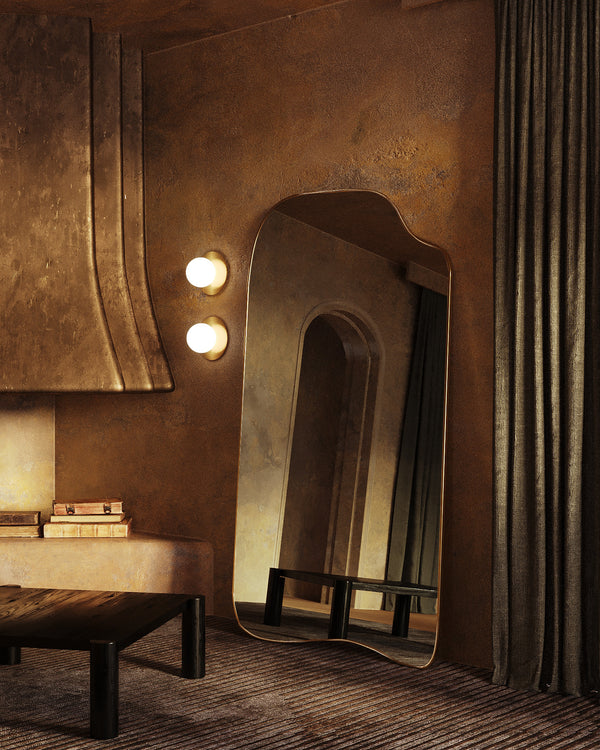 A full-length mirror with a softly curved frame leaning against a textured wall, styled in a warm, modern interior with wall lights, a low wooden bench, stacked books, and floor-length curtains.