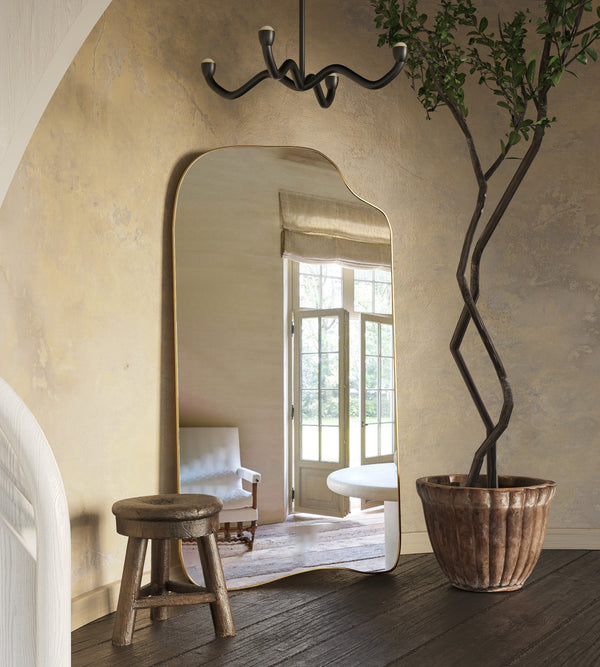 A sculptural full-length mirror with a slim brass frame leaning against a textured wall, reflecting a sunlit window, styled with a wooden stool, a potted tree, and modern ceiling lighting in a warm interior setting.