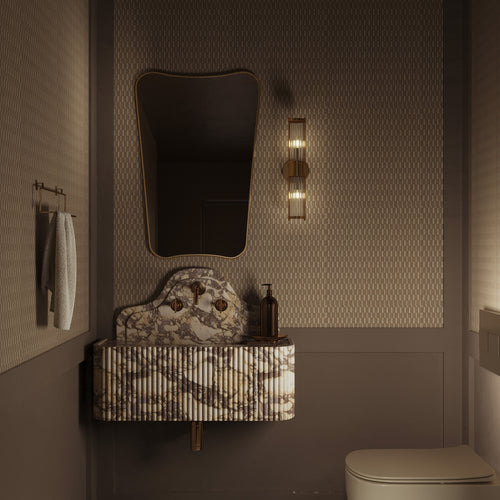 A bathroom vanity with a sculptural marble sink and fluted front, paired with an irregular-shaped mirror, brass wall sconce, patterned wallpaper.