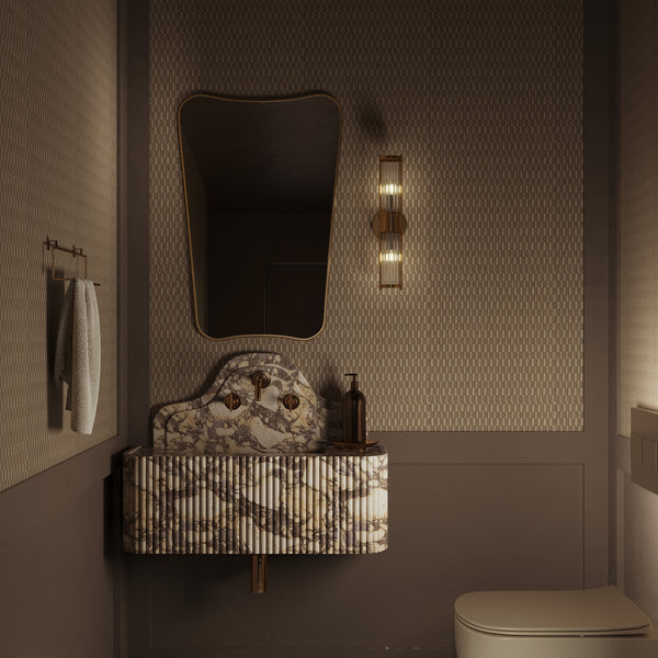 A bathroom vanity with a sculptural marble sink and fluted front, paired with an irregular-shaped mirror, brass wall sconce, patterned wallpaper.