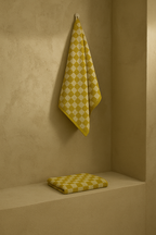 Yellow mustard and white checkered towel hanging on a wall hook, with a folded matching towel placed on a shelf below, highlighting the geometric pattern and terry texture.