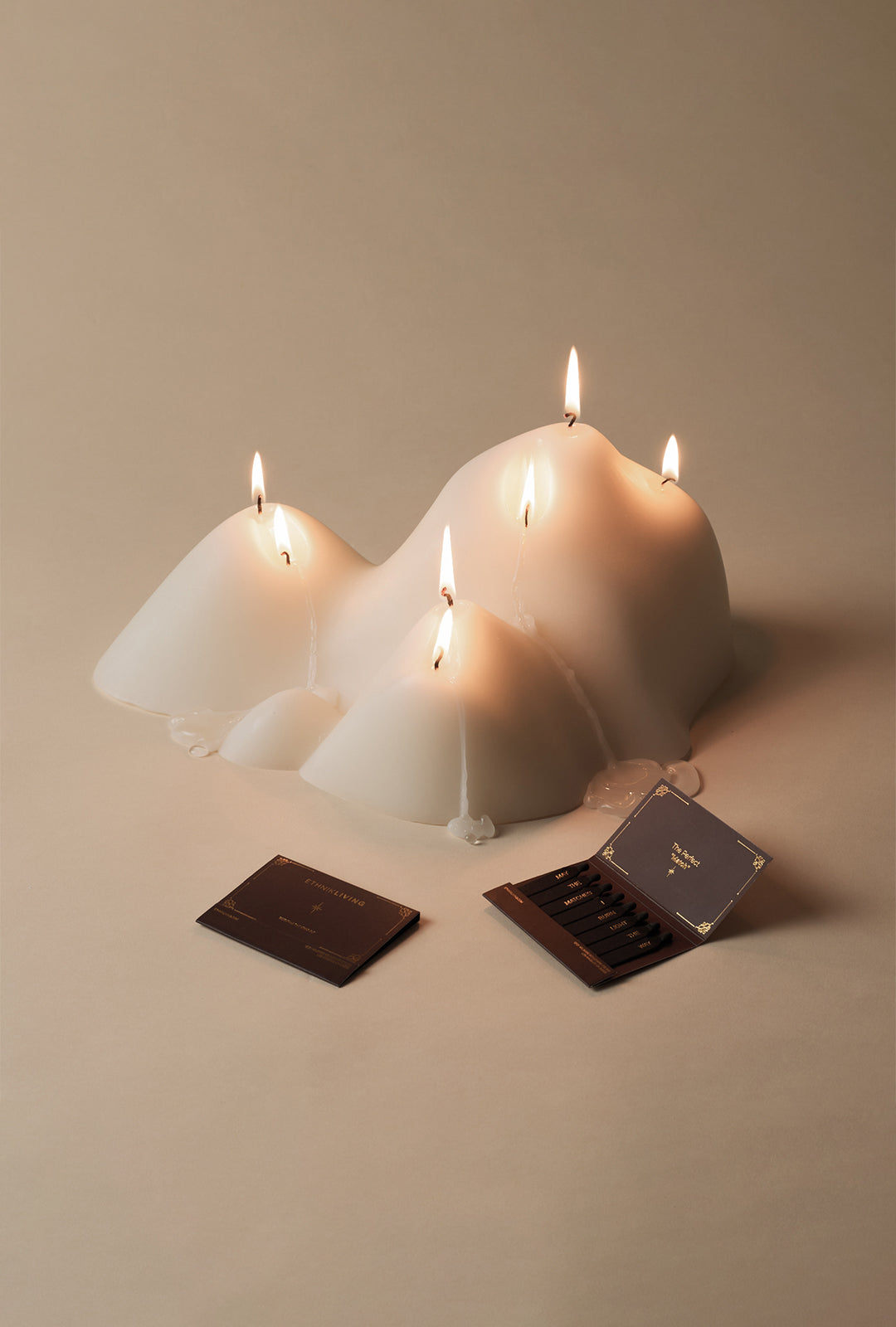 White sculptural candle with an organic, mountain-inspired form and multiple lit wicks, softly melting wax visible at the base. Two small matchbook packages are placed in front of the candle on a neutral surface against a warm, minimal background.