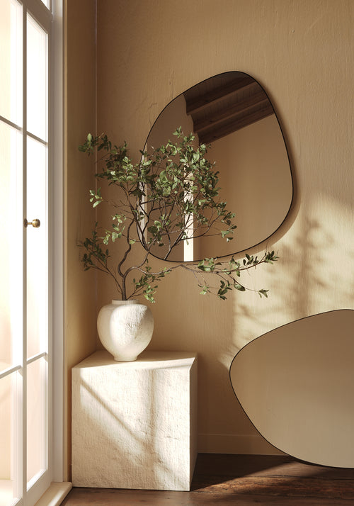 Organic-shaped wall mirror above a sculptural pedestal with a ceramic vase and leafy branches near a window.