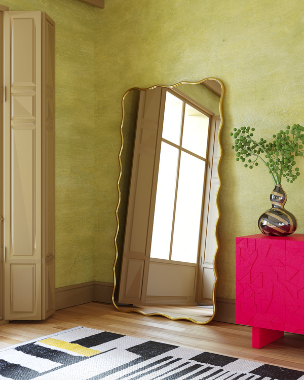 Wavy-edged full length mirror leaning against a green wall beside a bright pink cabinet with a glass vase.