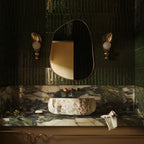 Asymmetrical brass-framed mirror mounted above a stone vessel sink in a bathroom with green tile walls and brass sconces.