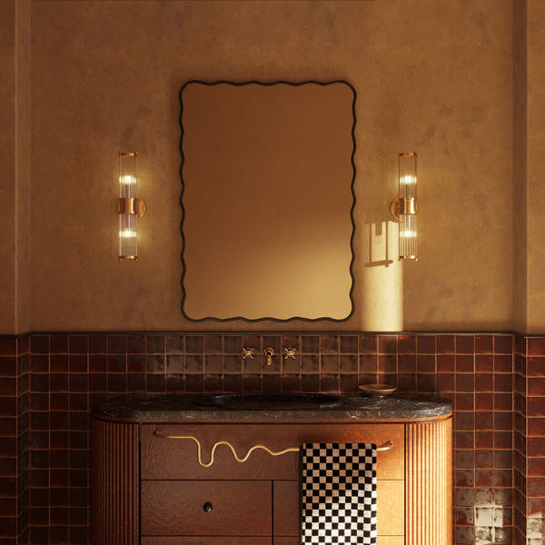 A warm-toned bathroom featuring a wavy-edged mirror, two wall-mounted sconces, a dark stone sink on a wooden vanity, and brown ceramic tile walls.