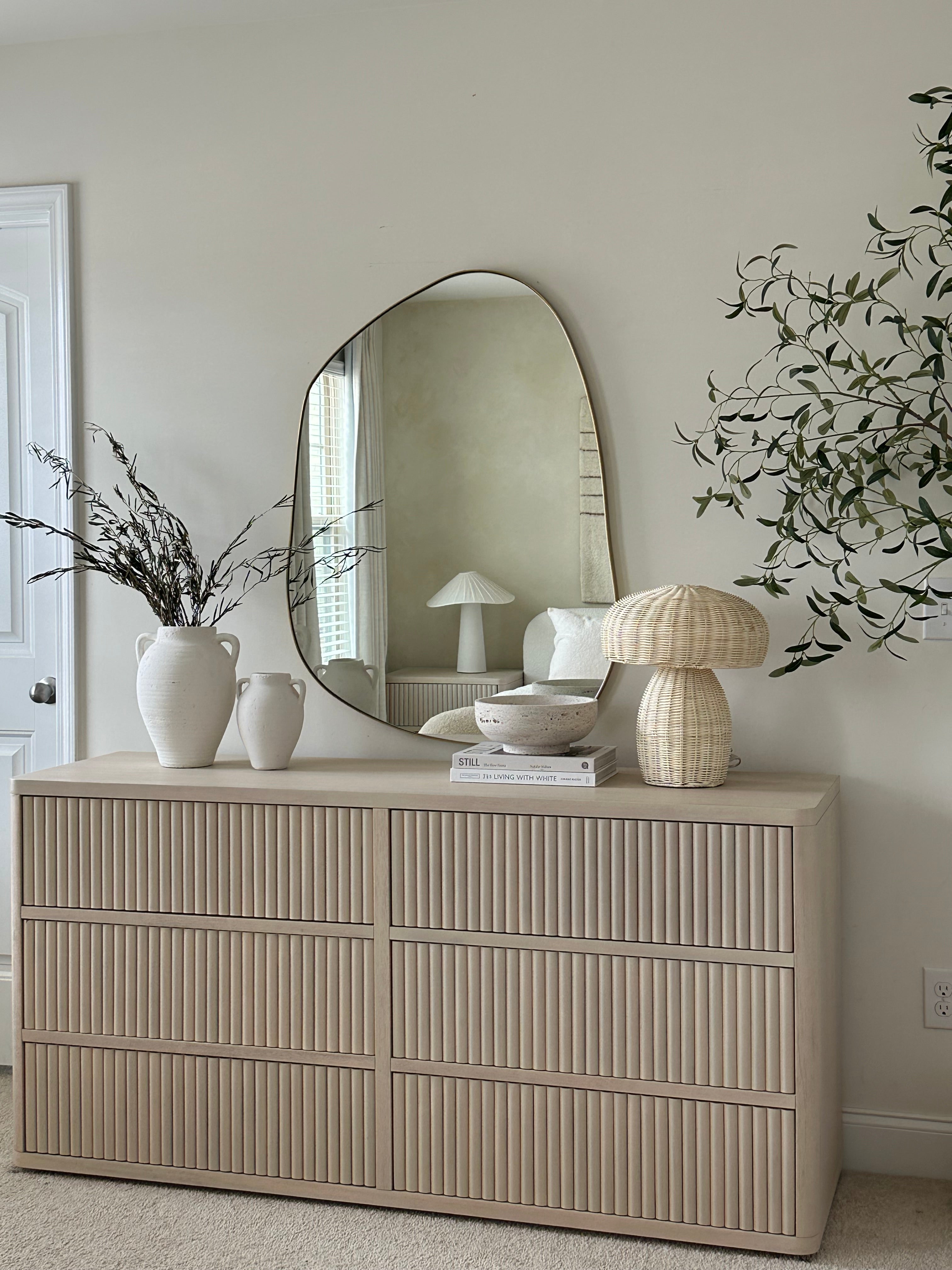 Wood dresser with vertical panel detailing, styled with decorative vases, books, a table lamp, and an irregular-shaped wall mirror.