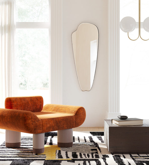 A sculptural wall mirror with an organic silhouette hangs between tall windows in a bright living room, styled above a patterned rug and next to an upholstered lounge chair.