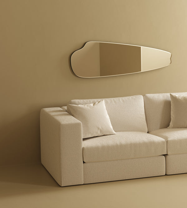 A minimalist living room with a beige modular sofa and a cream pillow, styled against a warm neutral wall with an elongated organic-shaped mirror mounted above.