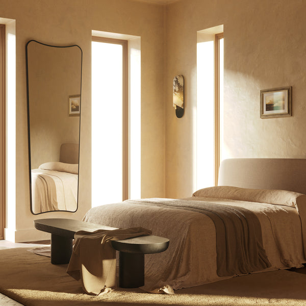 Minimalist bedroom with a low upholstered bed, textured area rug, dark wood bench, and a tall wavy-edge mirror reflecting soft natural light from narrow windows.