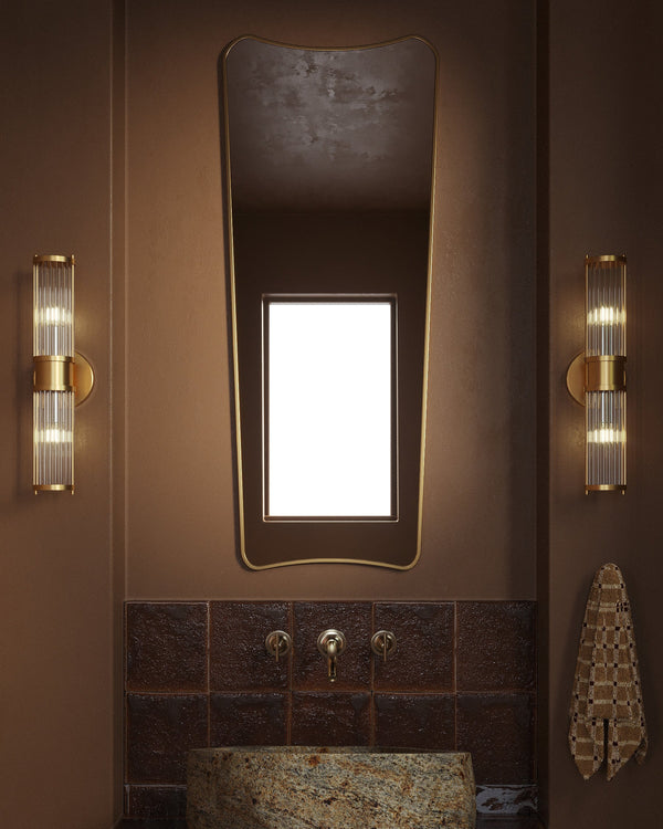 Full-length mirror with a softly curved, asymmetrical silhouette mounted above a bathroom sink, flanked by two wall lights in a warm-toned interior.