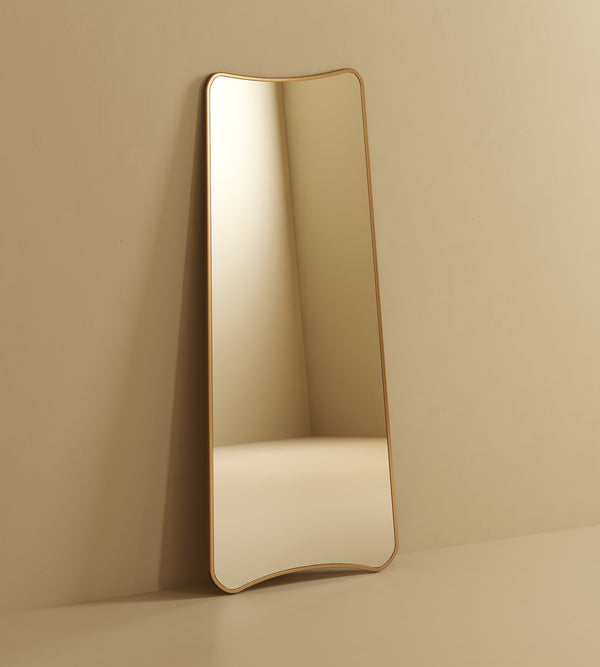 A full-length mirror with a soft, organic silhouette and slim gold frame leans against a neutral beige wall in a minimalist interior.
