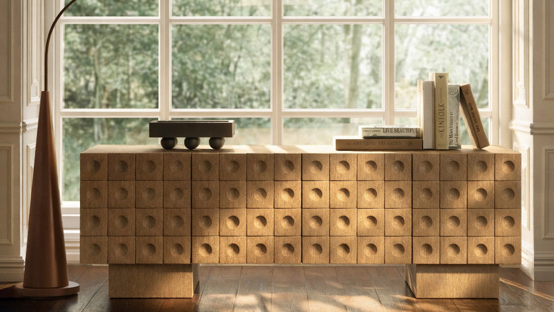 Textured wooden sideboard styled in front of large windows with books and a sculptural floor lamp.