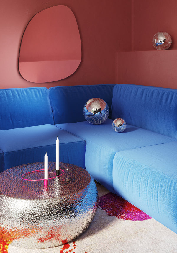 Blue sectional sofa in a pink room with an organic-shaped wall mirror, a silver round coffee table, and decorative disco ball accents.