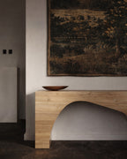 Wood console table with an arched base positioned against a wall beneath a large wall tapestry.