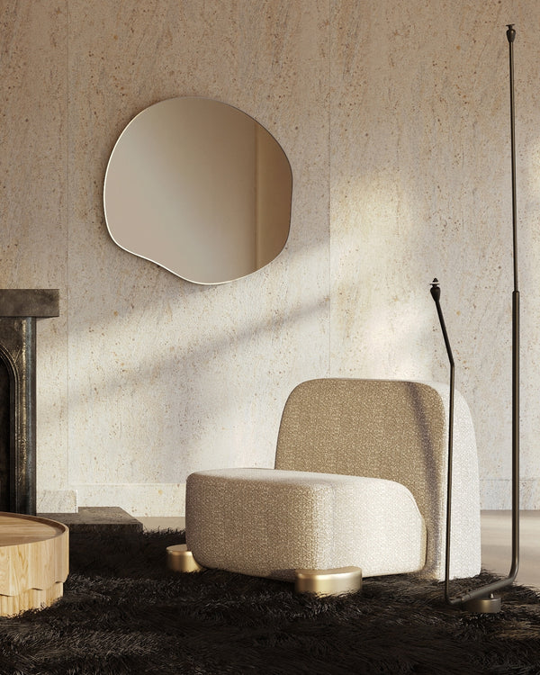 An organically shaped wall mirror is mounted on a textured stone wall above a sculptural lounge chair, styled in a minimalist living room with a black floor lamp and dark area rug.