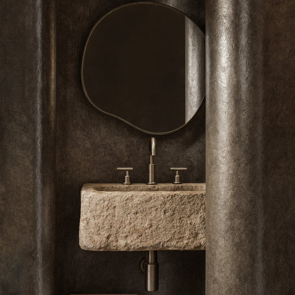 Organic-shaped wall mirror with a slim stainless steel frame mounted above a stone sink in a textured bathroom interior.