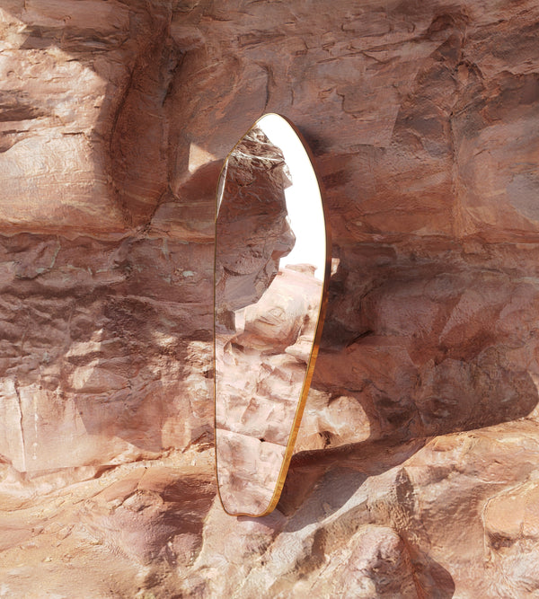 Asymmetrical brass-framed full length mirror placed against textured natural rock, reflecting the surrounding stone landscape.