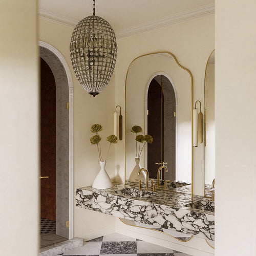An arched brass-framed mirror mounted above a marble bathroom vanity, styled with white ceramic vases, green floral stems, and brass fixtures in a softly lit, elegant bathroom.