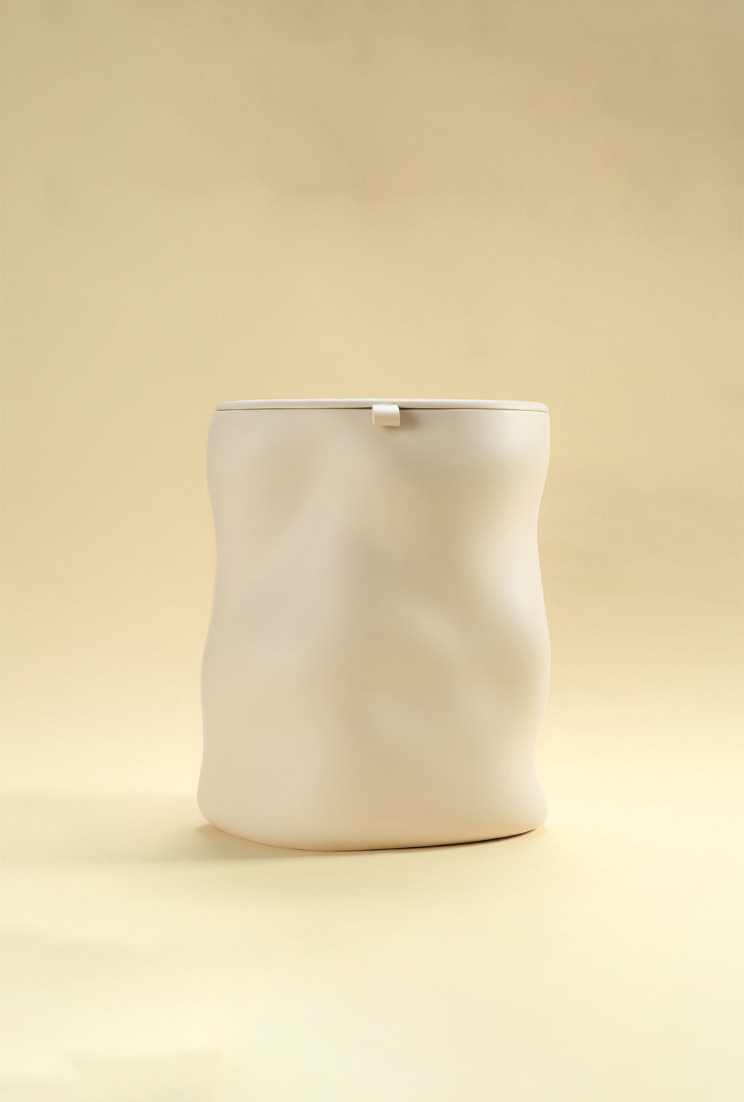 A beige sculptural dustbin with a fitted lid, photographed against a light yellow background.