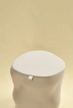 A beige sculptural dustbin with a fitted lid and a small front pull tab, photographed against a light yellow background.