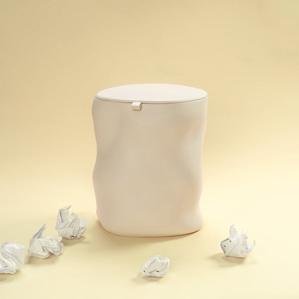 A beige sculptural dustbin with a fitted lid, surrounded by several crumpled pieces of white paper on the floor, against a light yellow background.