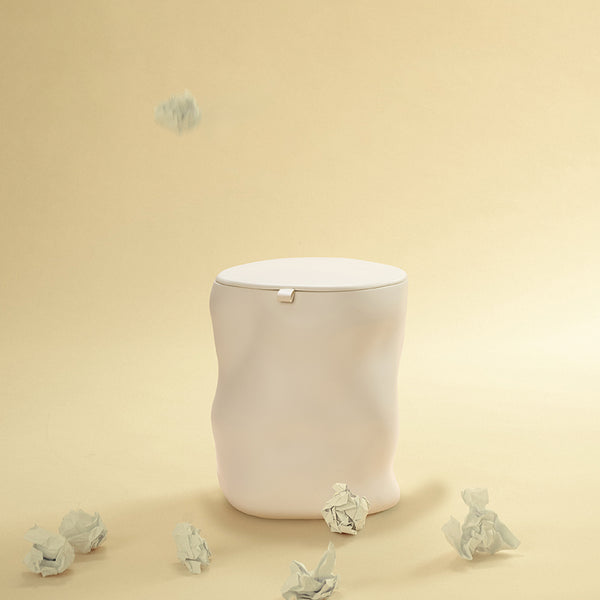A beige sculptural dustbin with a fitted lid, with several crumpled pieces of white paper on the floor around it and one piece suspended above the bin, against a light yellow background.