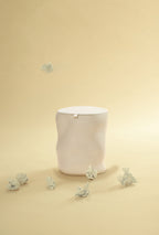 A beige sculptural dustbin with a fitted lid, with several crumpled pieces of white paper on the floor around it and one piece suspended above the bin, against a light yellow background.