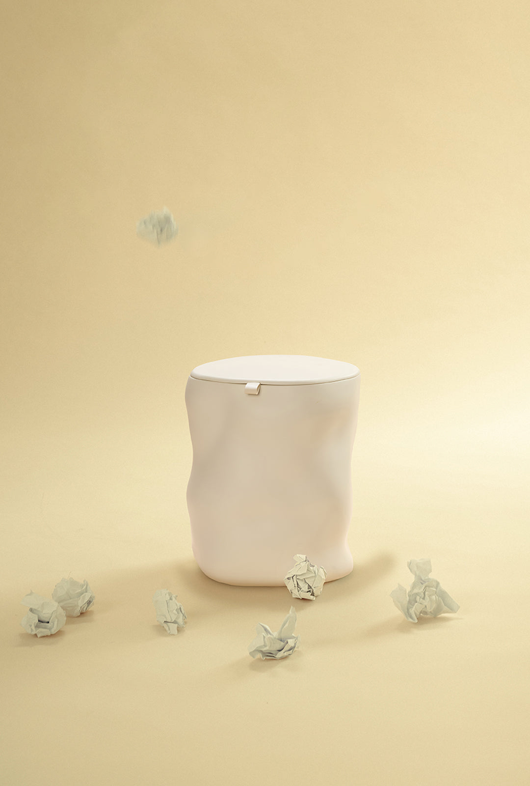 A beige sculptural dustbin with a fitted lid, with several crumpled pieces of white paper on the floor around it and one piece suspended above the bin, against a light yellow background.