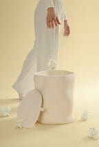 A beige sculptural dustbin with its lid placed to the side, as a person releases a crumpled piece of white paper into the open bin, against a light yellow background.