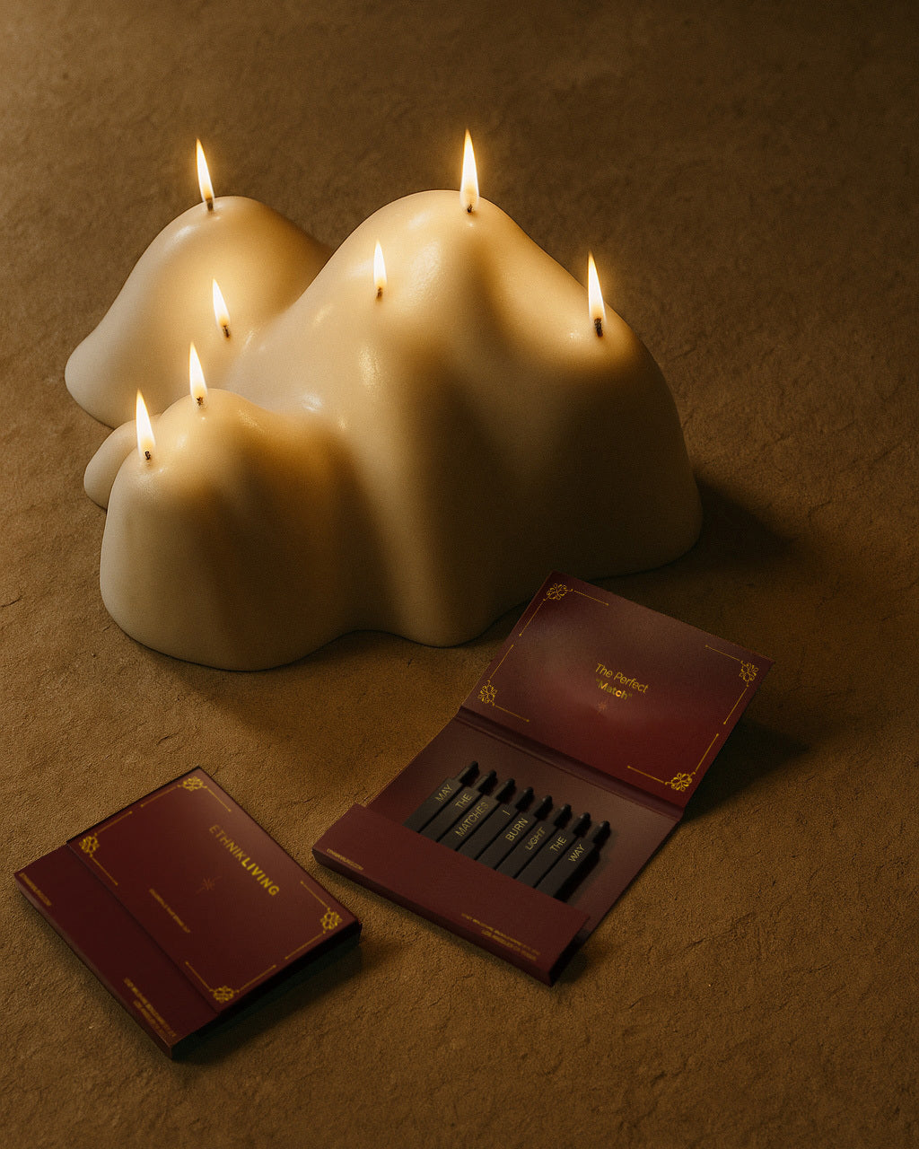 Mountain-shaped white candle with several lit wicks, styled beside open and closed matchboxes in a warm-toned setting.