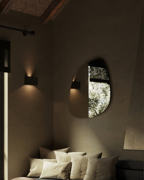 A softly shaped wall mirror mounted in a cozy interior corner, reflecting outdoor greenery, styled with neutral textiles, textured walls, and warm wall-mounted lighting.