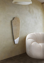 Frameless elongated mirror with an organic silhouette mounted on a textured wall beside a curved upholstered chair.