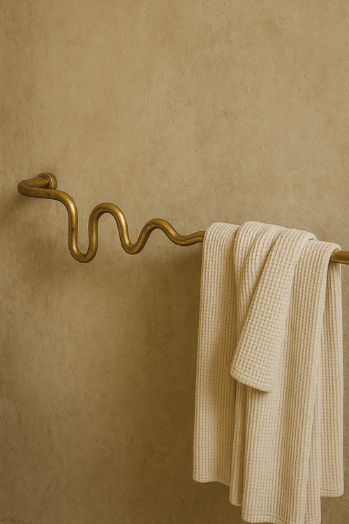 Textured neutral wall with a wavy gold brass towel bar mounted horizontally, holding a folded light-colored towel draped over one end.