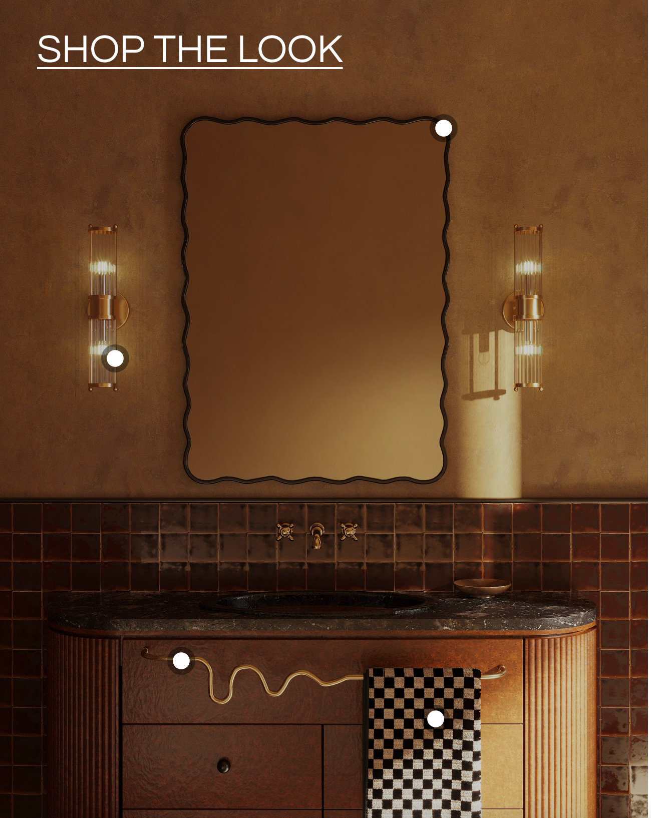A styled bathroom interior featuring a large rectangular wall mirror with a scalloped black frame, mounted above a dark wood vanity with a dark countertop. Two matching wall sconces illuminate the scene, and a checkered towel hangs on the cabinet.